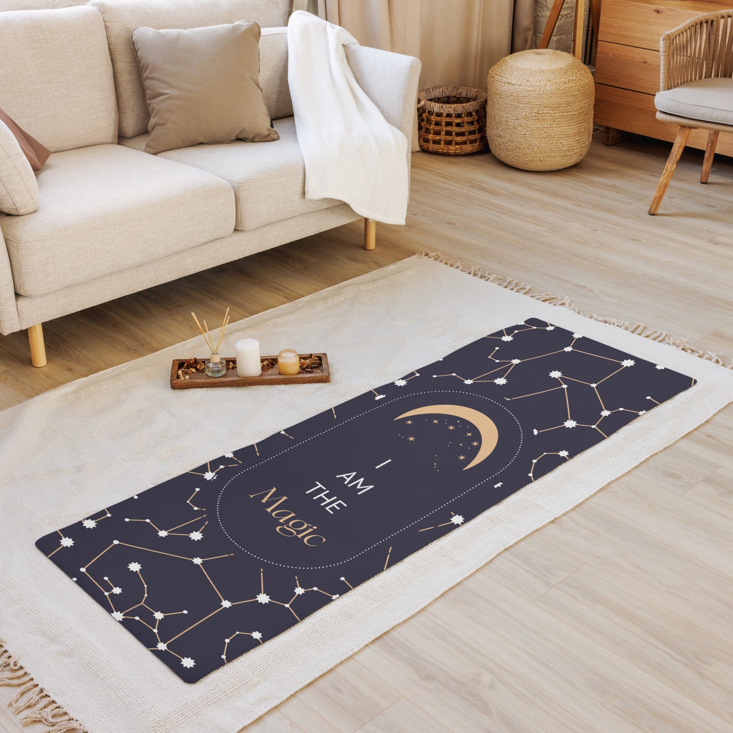 A yoga mat with a black and gray design featuring stars and the text 'I AM THE MAGIC'. The mat is placed on a wooden floor in a home setting, with a couch, chair, and candle holder in the background.