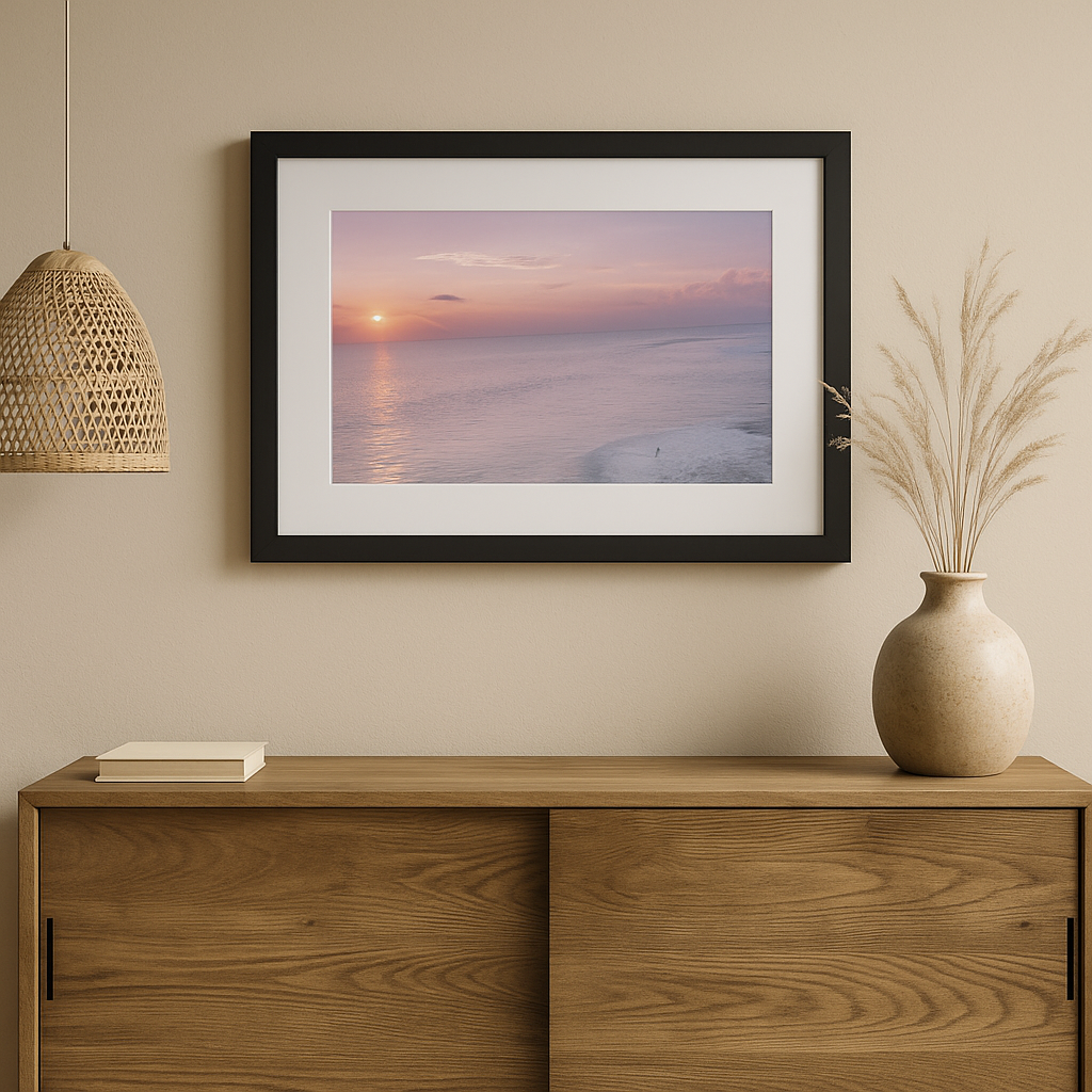 Framed artwork of a sunset over water on a beige wall above a wooden cabinet.