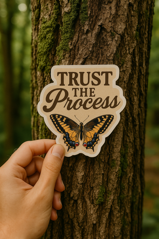 Hand holding a 'Trust the Process' sticker with a butterfly design against a tree background