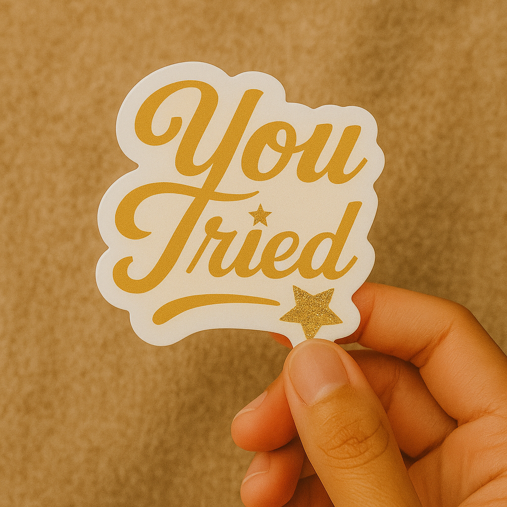 Hand holding a 'You Tried' sticker with gold text and star on a brown background